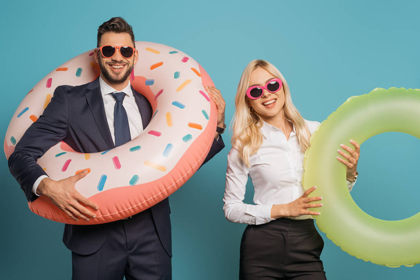 happy businesspeople in sunglasses holding inflatable rings while looking at camera on blue background - Photo, Image