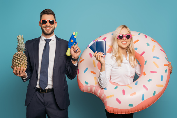 gelukkige zakenmensen met zwemring, documenten, waterpistool en verse ananas glimlachend op camera op blauwe achtergrond - Foto, afbeelding