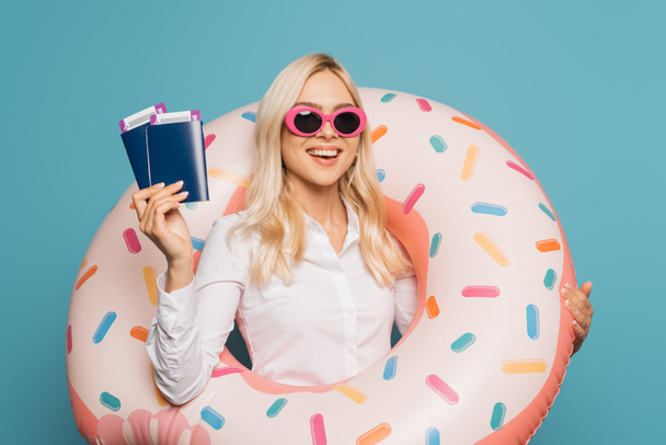 gelukkige zakenvrouw in zonnebril en zwemring met paspoorten en vliegtickets geïsoleerd op blauw - Foto, afbeelding