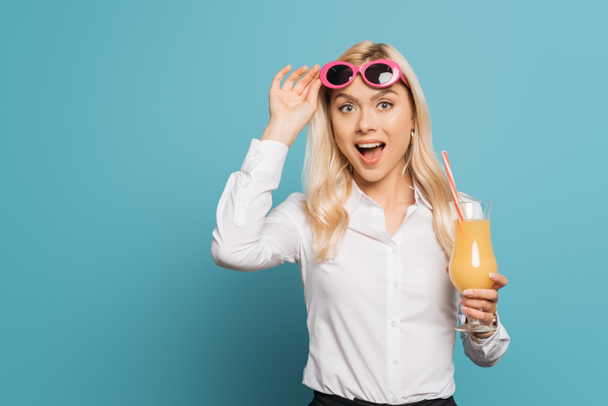 excited businesswoman touching sunglasses while holding orange juice and looking at camera on blue background - Photo, image