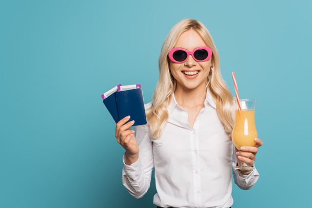 χαρούμενη επιχειρηματίας με γυαλιά ηλίου που κρατά ένα ποτήρι χυμό πορτοκαλιού, διαβατήρια και αεροπορικά εισιτήρια σε μπλε φόντο - Φωτογραφία, εικόνα