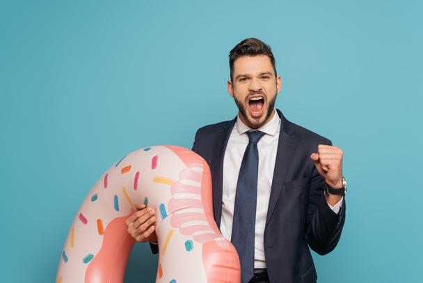 excited businessman showing yes gesture and shouting while holding inflatable ring on blue background - Фото, изображение