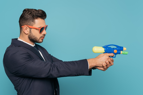 side view of businessman in sunglasses holding toy water gun isolated on blue - Foto, afbeelding