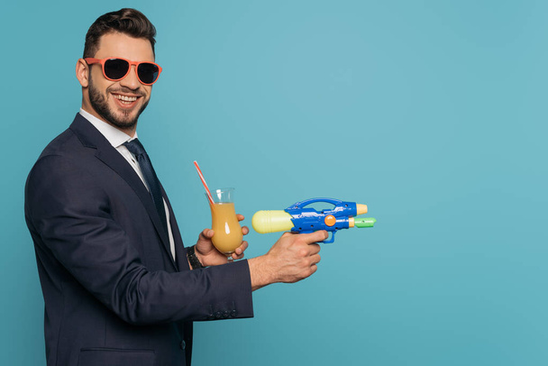 alegre empresario sosteniendo pistola de agua y vaso de jugo de naranja aislado en azul
 - Foto, imagen