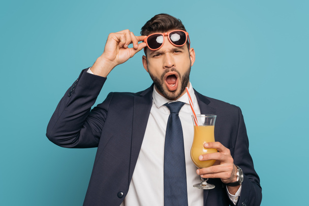 schockierter Geschäftsmann mit offenem Mund berührt Sonnenbrille, während er Orangensaft auf blauem Hintergrund hält - Foto, Bild