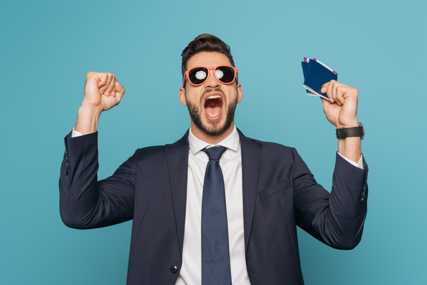 podekscytowany biznesmen w okularach przeciwsłonecznych krzyczy i pokazuje gest zwycięzcy trzymając paszporty i bilety lotnicze odizolowane na niebiesko - Zdjęcie, obraz