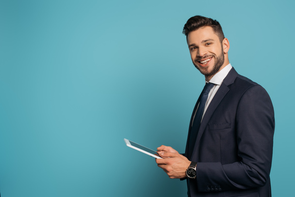 handsome businessman using digital tablet while smiling at camera on blue background - Photo, Image