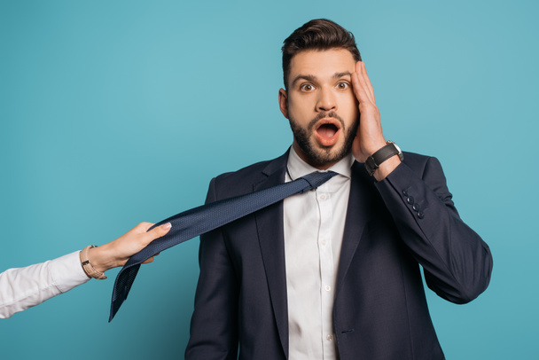 cropped view of woman pulling tie of shocked businessman touching face on blue background - Фото, зображення