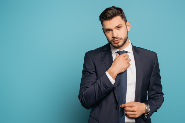 confident, handsome businessman touching tie while looking at camera on blue background - Foto, Bild