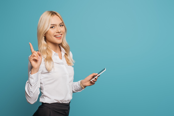 glückliche Geschäftsfrau zeigt Ideengeste, während sie ihr Smartphone in die Kamera hält und isoliert auf blau blickt - Foto, Bild