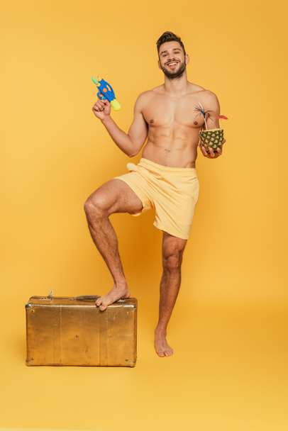 happy shirtless man holding water gun cocktail in pineapple while stepping on vintage suitcase on yellow background - 写真・画像