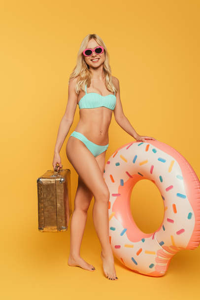 attractive girl in swimsuit and sunglasses holding swim ring and vintage suitcase on yellow background - Fotoğraf, Görsel