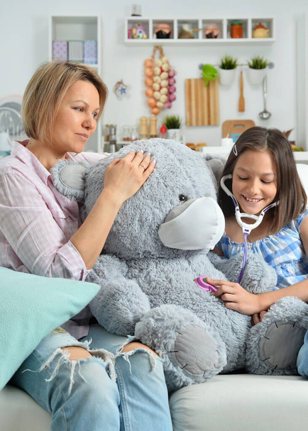  little girl and mother playing   with toy teddy  bear in facial mask. Girl with stethoscope - Zdjęcie, obraz