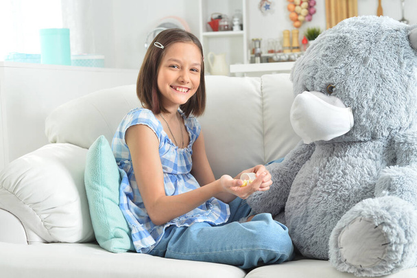   girl  giving pills to toy teddy  bear in facial mask on sofa - Фото, изображение