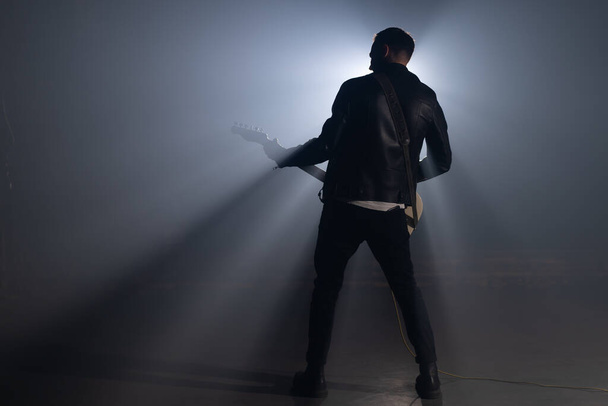Rock guitarist man in leather jacket in smoky studio or stage masterfully playing electric guitar. View of musician in the spotlight. - Fotó, kép