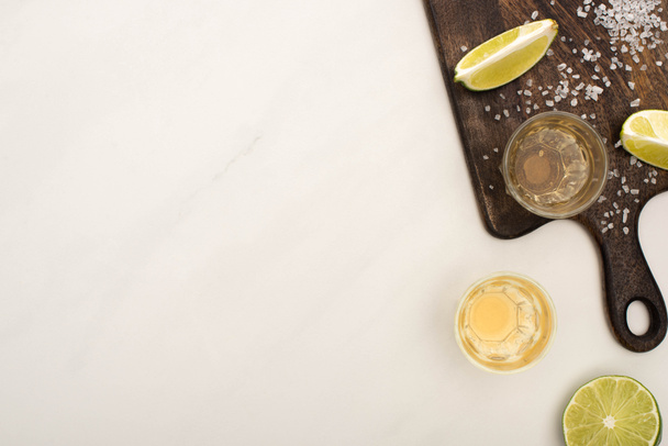 top view of golden tequila with lime, salt on wooden cutting board on white marble surface - Fotoğraf, Görsel