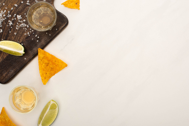 top view of golden tequila with lime, salt and nachos near wooden cutting board on white marble surface - Photo, Image