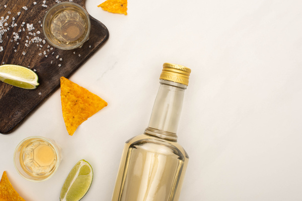 top view of golden tequila with lime, salt and nachos near wooden cutting board on white marble surface - Foto, imagen