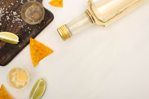 top view of golden tequila with lime, salt and nachos near wooden cutting board on white marble surface - Foto, immagini