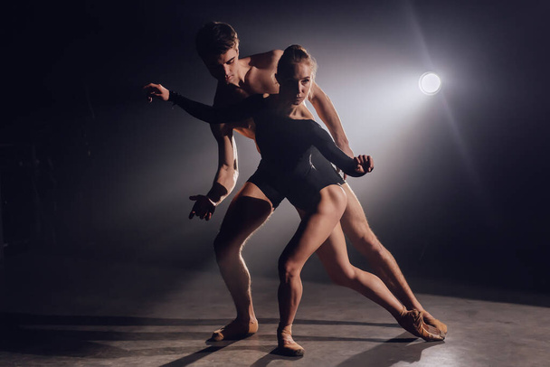 Beautiful young acrobats or gymnasts on floodlights background. Professional ballet couple dancing in spotlights smoke on big stage. Emotional duet performing choreographic art - Фото, зображення