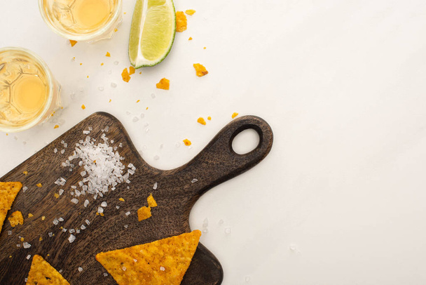 vrchní pohled na zlatou tequilu v broušených sklenicích s vápnem, solí a nachos na dřevěné řezací desce na bílém mramorovém povrchu - Fotografie, Obrázek