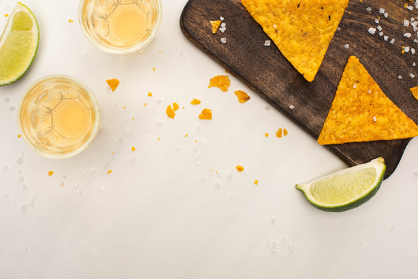 top view of golden tequila in shot glasses with lime, salt and nachos on wooden cutting board on white marble surface - Foto, afbeelding