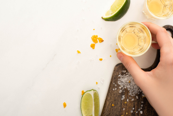 bijgesneden beeld van vrouw drinken gouden tequila met limoen, zout op wit marmeren oppervlak - Foto, afbeelding