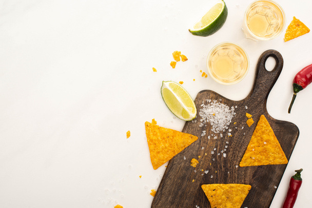 top view of golden tequila in shot glasses with chili peppers, lime, salt and nachos on wooden cutting board on white marble surface - Foto, Imagem