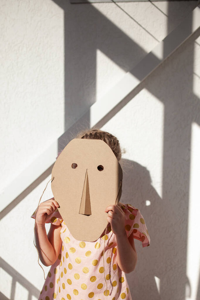 Portret van een meisje. Een kind verbergt zijn gezicht achter een kartonnen masker, binnen - Foto, afbeelding