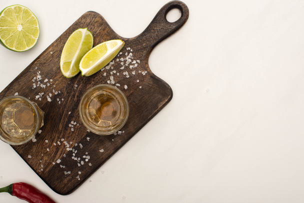 top view of golden tequila with lime, chili pepper, salt on wooden cutting board on white marble surface - Valokuva, kuva