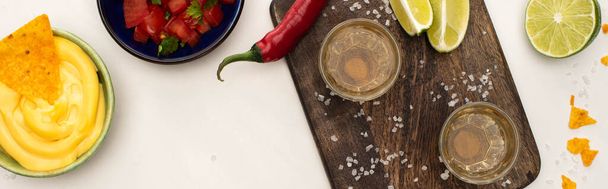 top view of golden tequila with lime, chili pepper, salt and nachos with cheese sauce near wooden cutting board on white marble surface, panoramic shot - Photo, Image