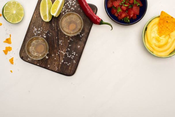 top view of golden tequila with lime, chili pepper, salt and nachos with cheese sauce near wooden cutting board on white marble surface - Фото, изображение
