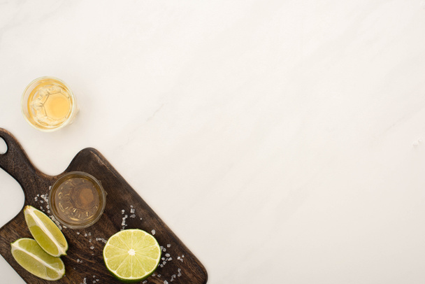 top view of golden tequila with lime, salt on wooden cutting board on white marble surface - Foto, immagini