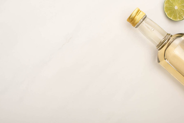 bovenaanzicht van gouden tequila in fles met kalk op wit marmeren oppervlak - Foto, afbeelding