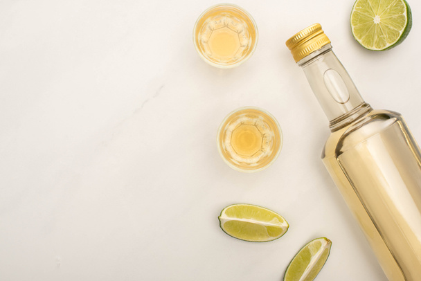 top view of golden tequila in bottle and shot glasses with lime on white marble surface - 写真・画像