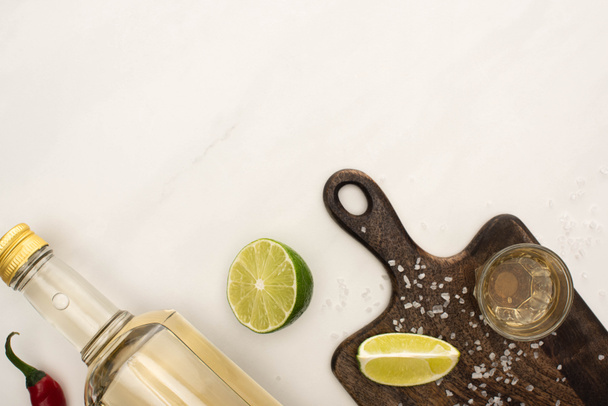 top view of golden tequila with lime, chili pepper, salt on wooden cutting board on white marble surface - Fotó, kép