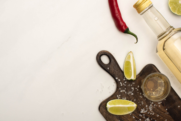 top view of golden tequila with lime, chili pepper, salt on wooden cutting board on white marble surface - Photo, Image