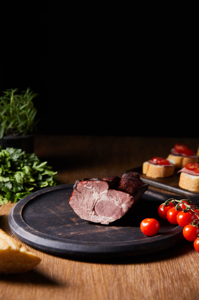 selective focus of tasty ham on board near canape, parsley, cherry tomatoes and baguette on wooden table isolated on black - Foto, afbeelding