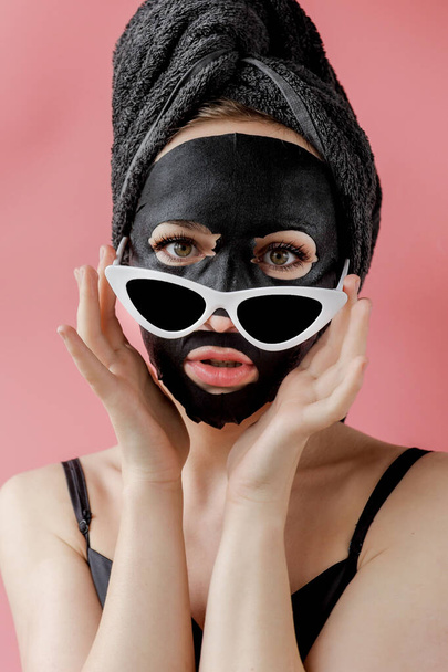 Young woman in glasses appling black cosmetic fabric facial mask on pink background. Face peeling mask with charcoal, spa beauty treatment, skincare, cosmetology. Close up. - Valokuva, kuva