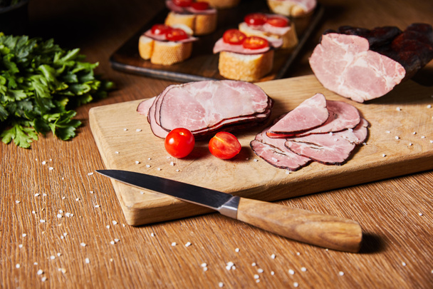 selective focus of tasty ham slices, cherry tomatoes and knife on cutting board near parsley and canape - Foto, Imagen