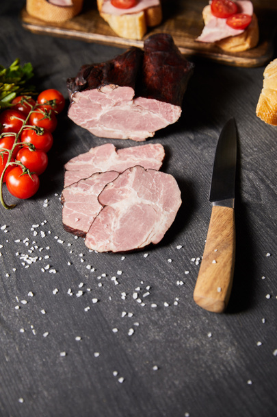selective focus of tasty ham sliced ham, cherry tomatoes, parsley, salt, knife and baguette on wooden grey table with canape - Photo, Image