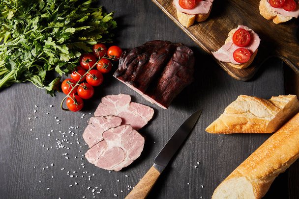 top view of tasty ham sliced ham, cherry tomatoes, parsley, salt, knife and baguette on wooden grey table with canape - Photo, image