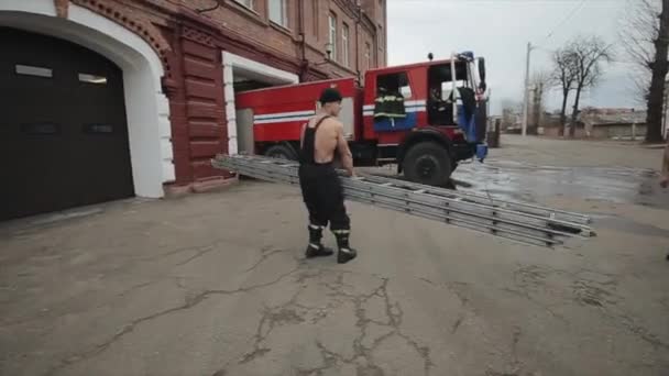 Νεαρός μυώδης άνδρας στρίβει μια μεγάλη μεταλλική σκάλα στα χέρια του στο φόντο ενός πυροσβεστικού φορτηγού. - Πλάνα, βίντεο