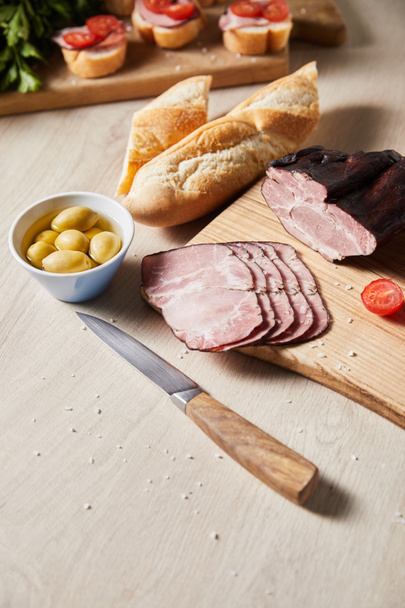 selective focus of tasty ham on cutting board with knife, olives and baguette on wooden table - Fotografie, Obrázek