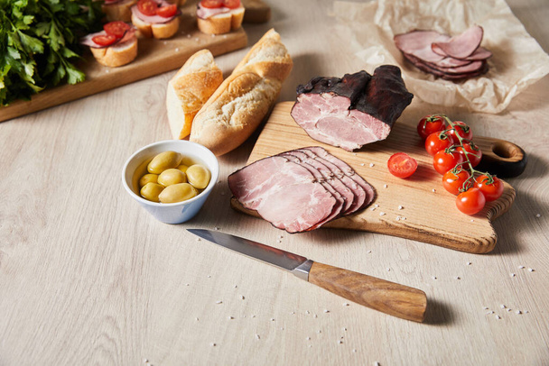 selective focus of tasty ham on cutting board with knife, parsley, cherry tomatoes, olives and baguette near canape on wooden table - Foto, afbeelding