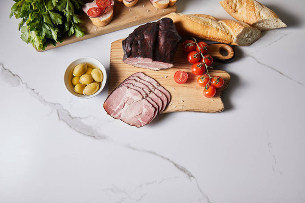 top view of tasty ham on cutting board with parsley, cherry tomatoes, olives and baguette on white marble surface - Φωτογραφία, εικόνα