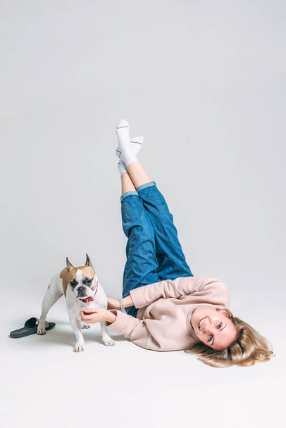 η νεαρή κοπέλα με το ροζ πουλόβερ βρίσκεται στο πάτωμα του στούντιο με λευκό φόντο και παίζει με το αστείο γαλλικό μπουλντόγκ της - Φωτογραφία, εικόνα