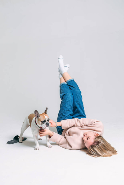 the young girl in a pink sweater lies on the floor in the studio with a white background and plays with her funny french bulldog - Foto, Imagen