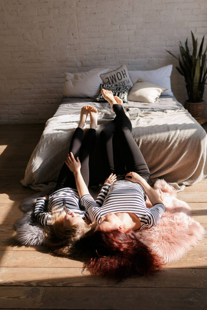 Little beautiful girl with long hair lies with her mother on the floor near the bed. Mom and daughter look around. They are dressed in striped clothes. Family at home. Loft style house. Mother's day concept. Selective focus. Medium sharp - 写真・画像