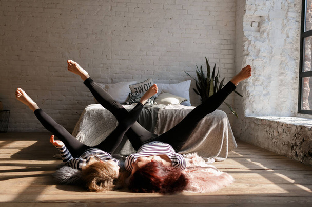 Little beautiful girl with long hair lies with her mother on the floor near the bed. Mom and daughter play sports together in Sunlight. They are dressed in striped clothes. Family at home. Loft style house. Mother's day concept. Selective focus. Medi - Valokuva, kuva